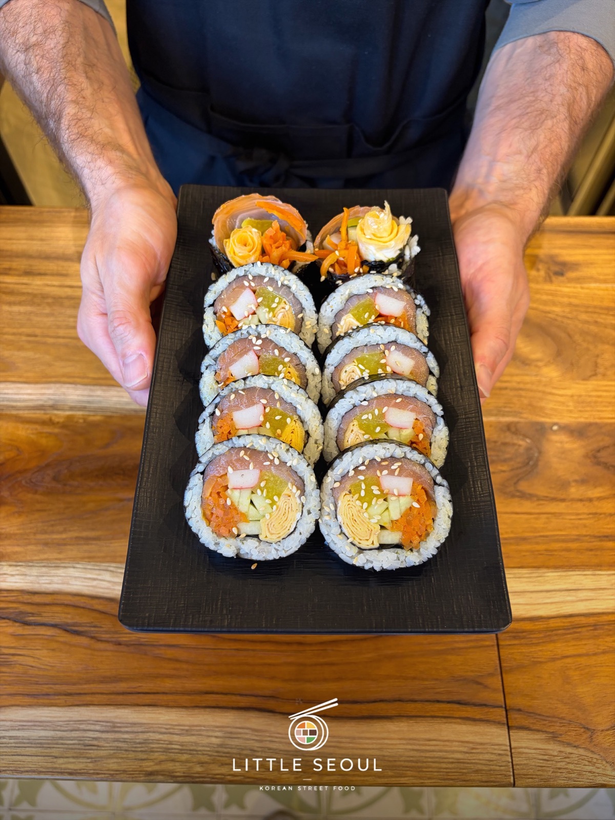 Hände reichen eine Platte mit frisch geschnittenem Kimbap über den Holztisch